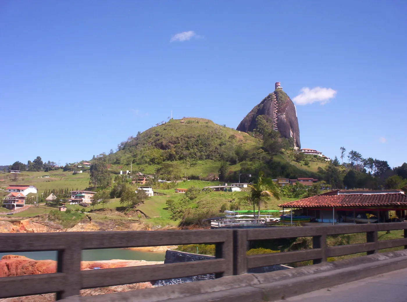 La Piedra del Peñol vista desde la carretera, con potreros y un barandal en primer plano